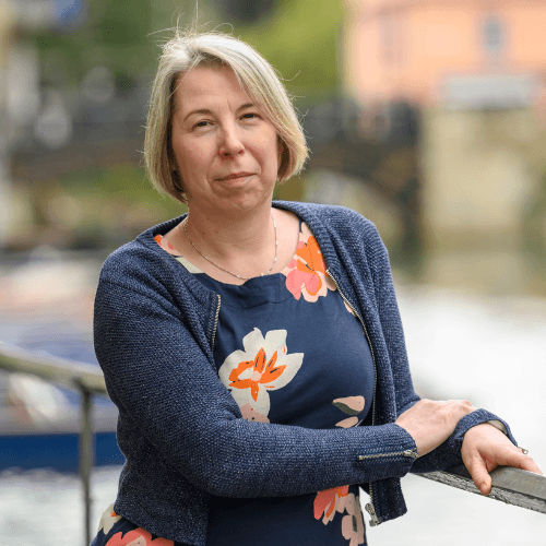 Emma, lady with blond hair wearing a dark blue top stands smiling in front of the river