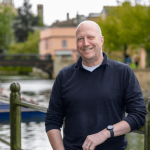 Mark, man wearing w dark blue polo shirt stands smiling in front of the river