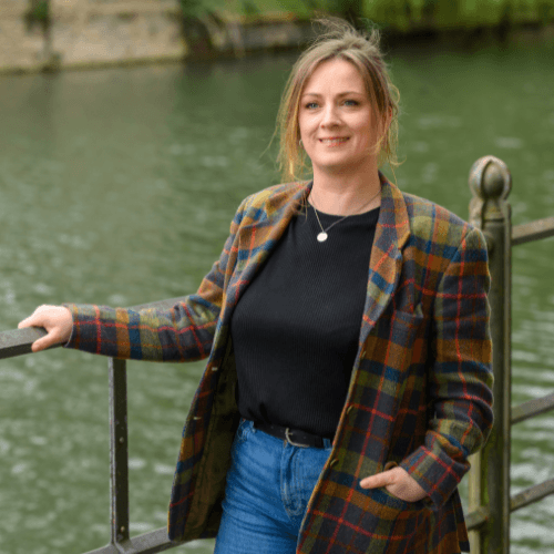 Trudy, lady with brown hair wearing a green, blue and red checkered coat stands smiling in front of the river