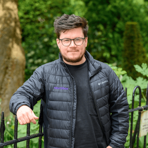 Andy - man wearing a black puffer jacket and glasses, stands smiling in front of a railing in a park