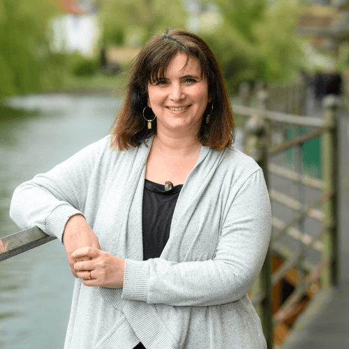 Dr Deb Leveoy - a lady with short brown hair standing against a rail smiling. She is wearing a waterfall light blue cardigan
