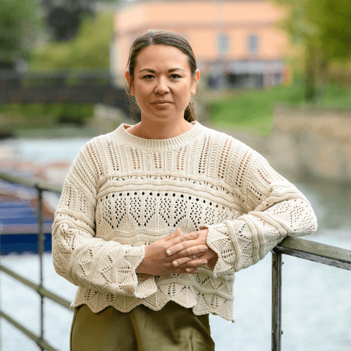 Jen Watton - a female in a white knit jumper stands smiling in front of the river