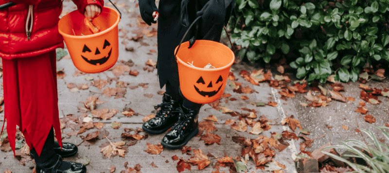 Two children trick or treating. One is wearing a red puffer jacket and red trousers carrying a pumpkin bucket. The other is wearing a black costume carrying a pumpkin bucket