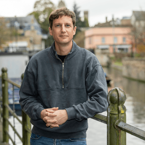 Ben Lewis, a white male with blonde hair in a grey jumper standing against a railing in front of a river