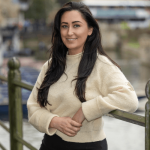 Jess Man - a female with dark brown long hair wearing a white jumper. jess is standing in front of the river against railings