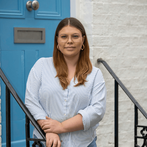 Chloe - a female in a blue shirt standing in front of a blue door. Chloe has shoulder length brown hair and is wearing glasses
