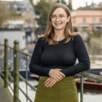 Bronwyn - they have short light brown hair and glasses. They are wearing a black longsleeved top and green trousers.