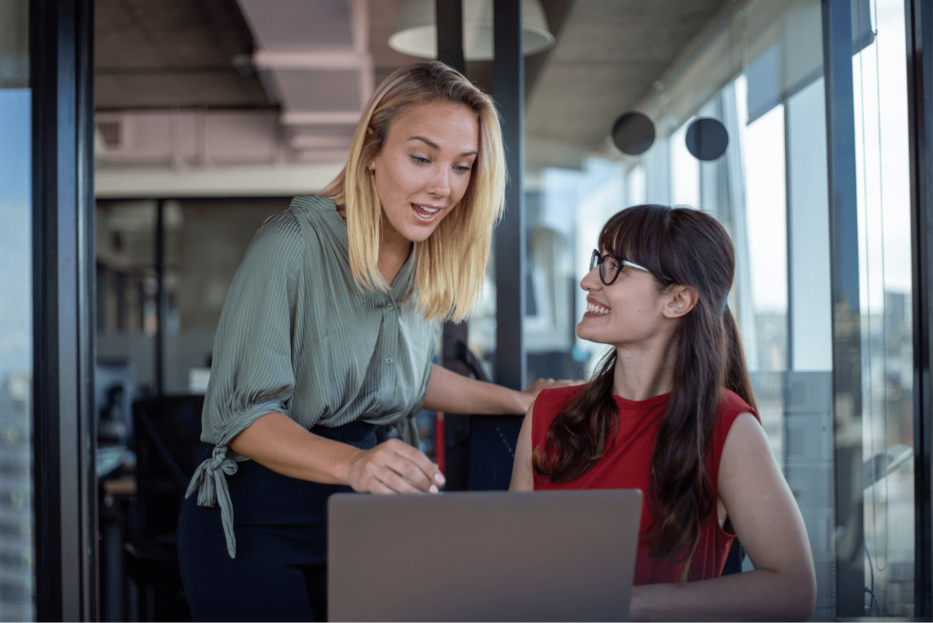 A female colleague, with blonde hair praising another female colleague with brunette hair, they are sitting around a computer screen