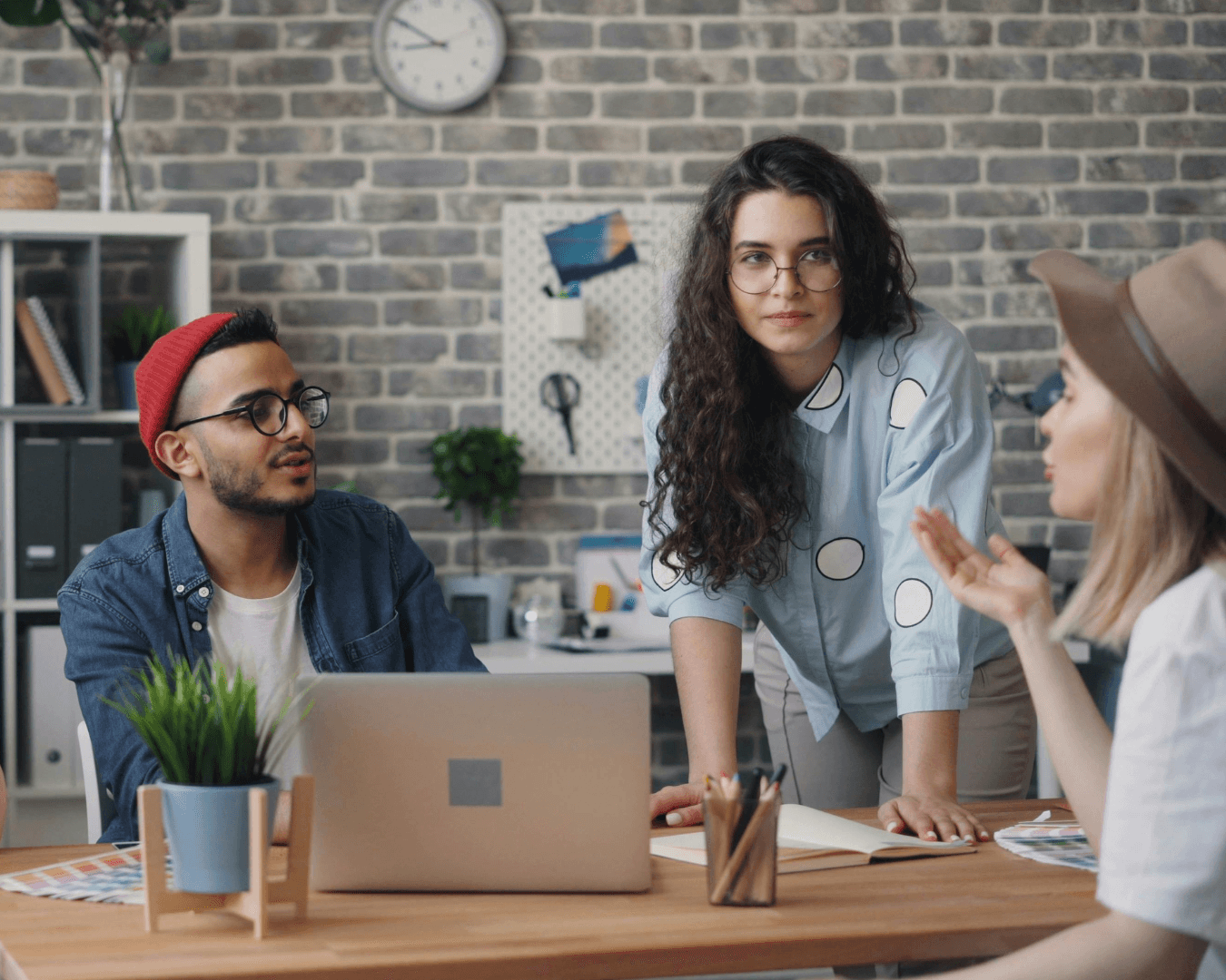 Two females, one blonde and one brunette, discussing work at a desk with a tanned male wearing a bright red hat