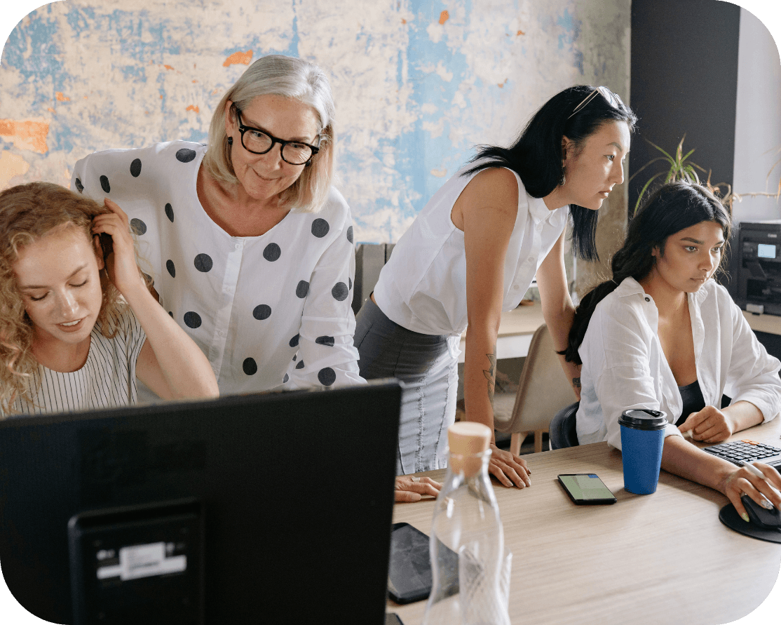 Two women standing with two female workers, assisting them on computers