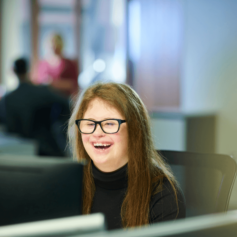 Smiling person with long brown hair and black glasses seated in an office meeting room, speaking while others sit blurred in the background; soft daylight and computer desks frame the scene.