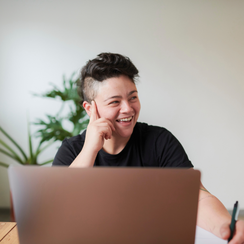 Person smiling and talking on a phone while working at a desk with a laptop, indoors with a potted plant in the softly blurred background.