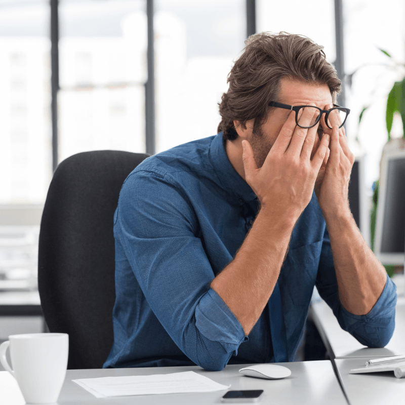 A person in a blue shirt sits at an office desk with papers, a smartphone, and a cup, covering their face with hands while wearing glasses, suggesting stress or frustration.